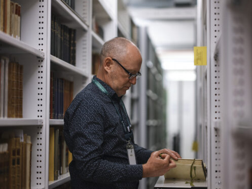 A man browsing the stacks of a library, holding an old book.